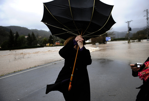 A woman attempts to hold onto her umbrella at a road partially submerged from floodwaters in Quinzanas, near Oviedo