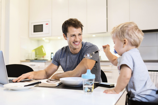 Happy father looking at baby boy while using laptop in kitchen
